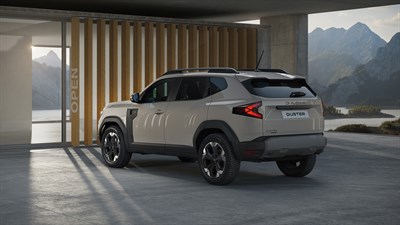 SUV Dacia Duster паркиран пред модерна куќа со планински пејзаж во позадина, беж боја на каросеријата и елегантен дизајн.