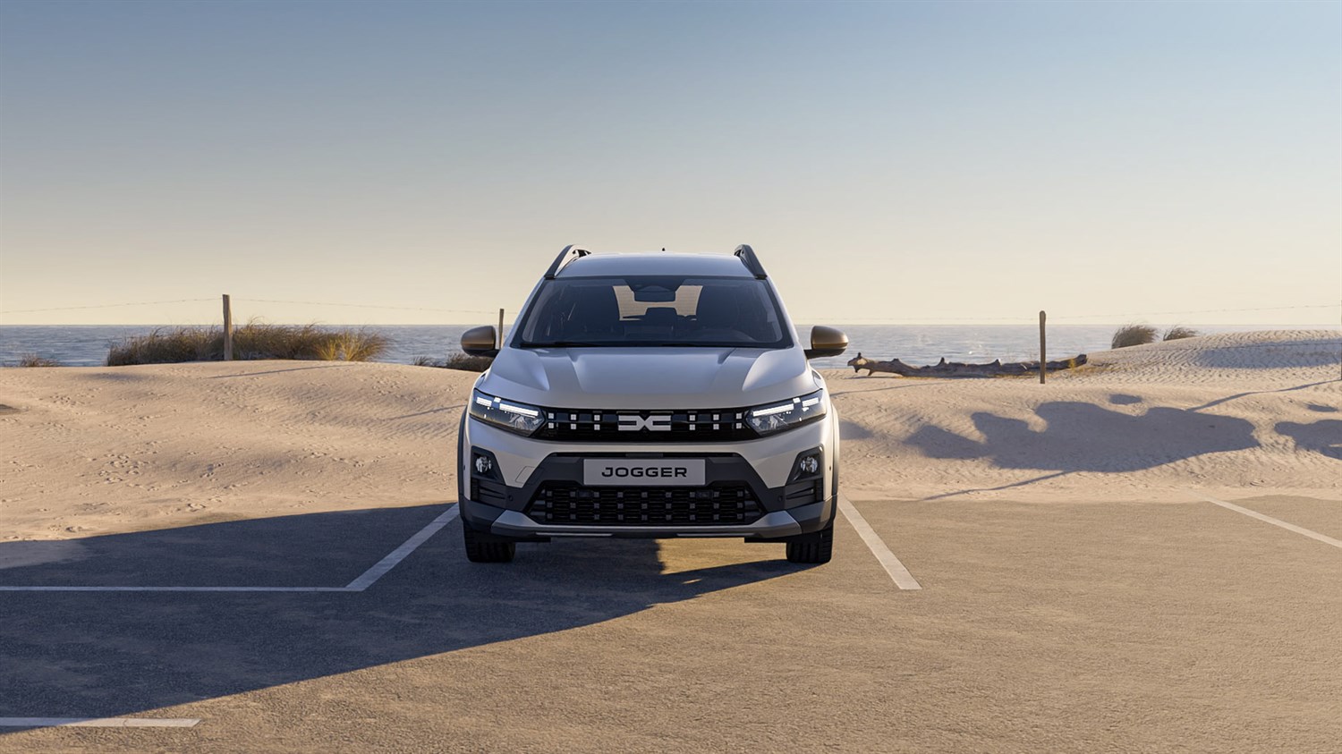 Dacia Jogger снимен од напред на паркинг покрај песочна плажа и морето.