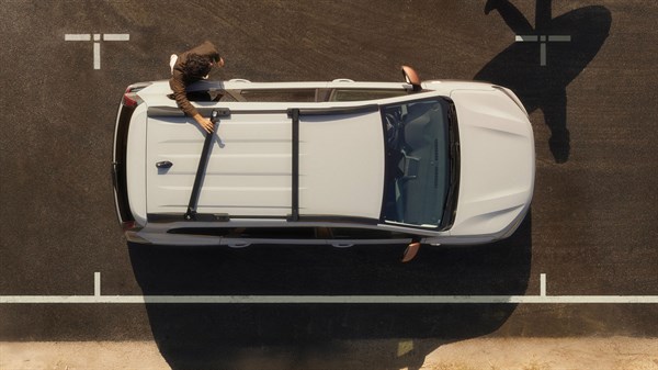 Dacia Jogger фотографиран одозгора на паркинг со држачи на кров и лице кое стои до возилото.