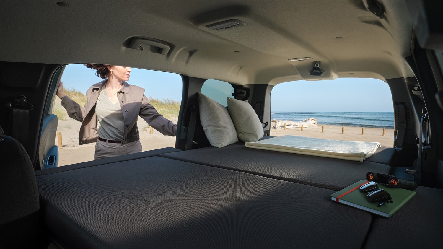 Внатрешноста на Dacia Jogger со целосно склопени задни седишта за да се создаде рамна површина слична на кревет, со перници и тетратка на задниот дел, и поглед на плажата низ отворените задни врати.