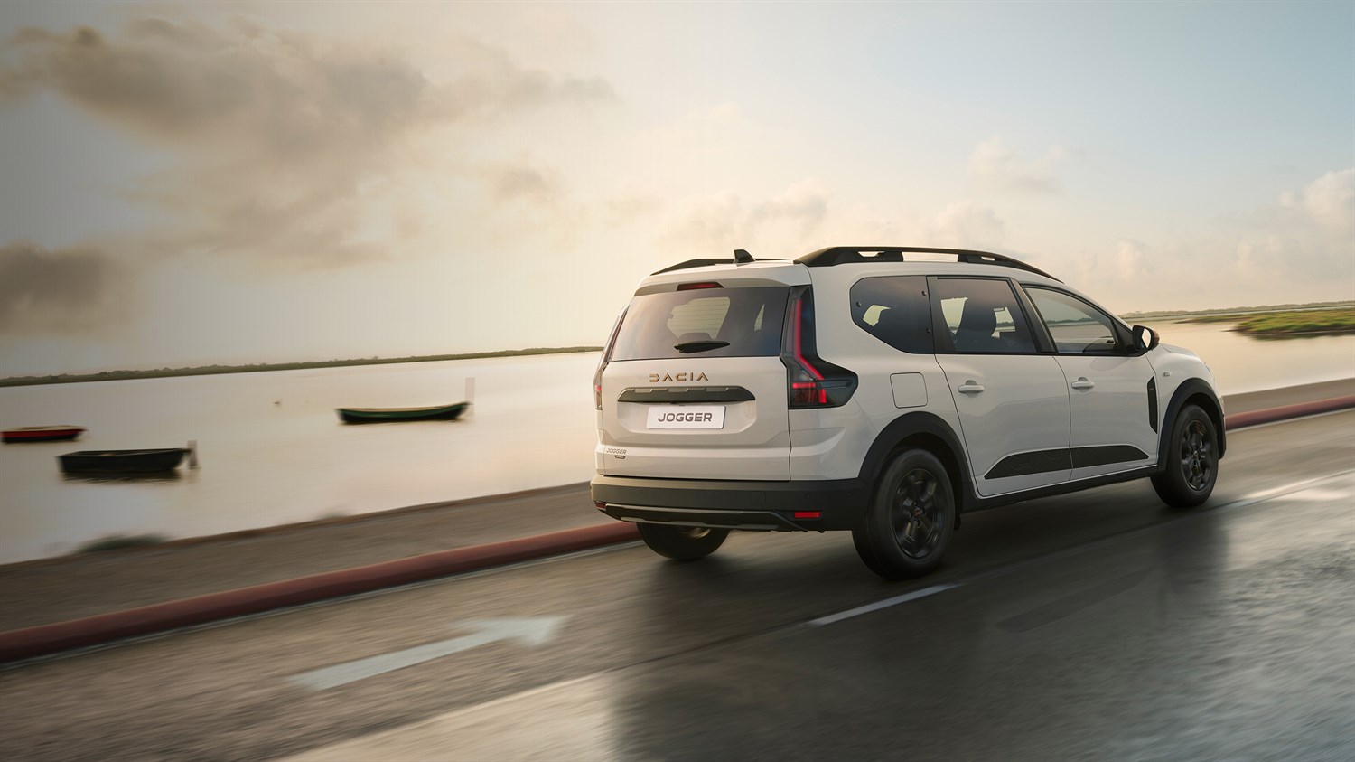 Бел Dacia Jogger прикажан одназад, вози по брегот, на пат со мирно море и бродови во позадина.