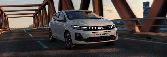 Возило Dacia Sandero вози преку модерен мост на зајдисонце, прикажано од напред со неговата препознатлива решетка и LED светла.