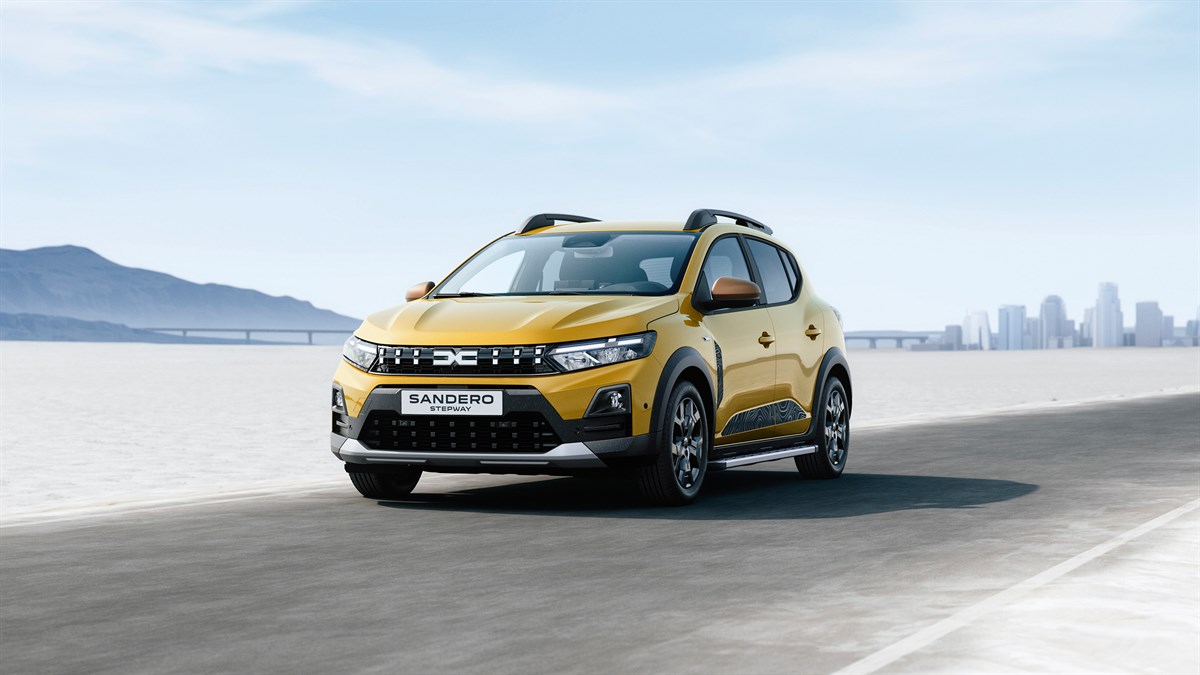 Dacia Sandero Stepway во жолта боја, фотографиран од напред додека возите низ пустински пејзаж, со планини и градска силуета во позадина.