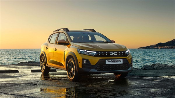 Dacia Sandero во жолта боја паркирано покрај морето на зајдисонце, фотографирано од напред лево.