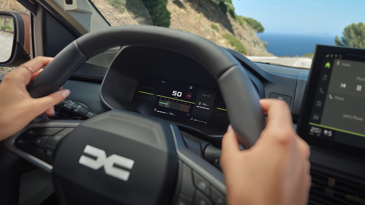 Возачот го држи управувачот на Dacia Sandero Stepway, со дигитален дисплеј за инструменти и поглед на крајбрежниот пат низ ветробранското стакло.